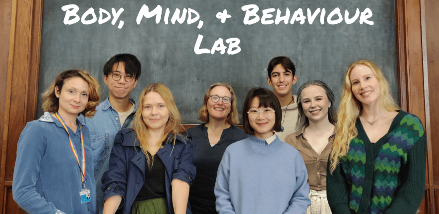 The Body, Mind, and Behaviour Laboratory members standing in front of a chalk board with their lab's name written in a chalk-style font. Members of the lab, from left to right, are Jasmina, Bryan, Elzė, Simone, Ekim, Edoardo, Veronice, and Katherine